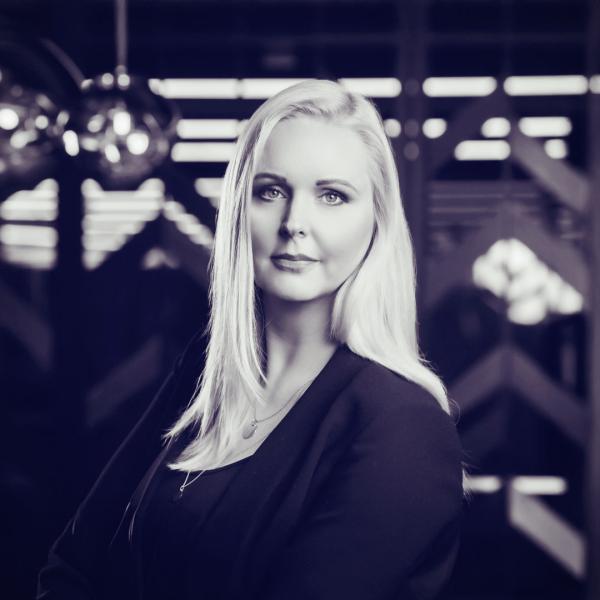 A photo of Gajzler Agnieszka, Managing Director TV&Digital Content Growth, Wirtualna Polska Group. A confident woman with long, straight blonde hair poses in a professional setting. She wears a dark blazer and a delicate necklace. The background features modern geometric patterns and pendant lights, creating a sophisticated atmosphere.