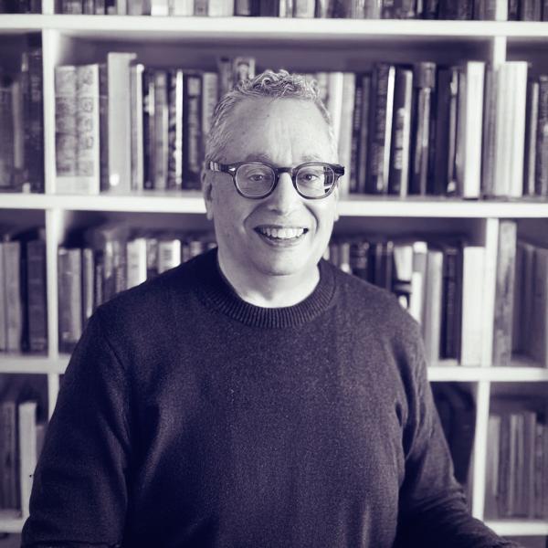 Black-and-white photo of Stuart Feil, an Independent Branded Content and Thought Leadership Consultant. He is smiling, wearing glasses and a dark sweater, standing in front of a bookshelf filled with books. The image has a slight purple tint.