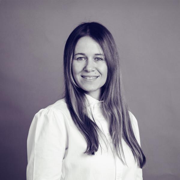 A professional black-and-white headshot of Ana Tomić. She has long, straight hair and is wearing a white button-down shirt. She is smiling warmly, exuding confidence and approachability, with a neutral background enhancing the focus on her face.