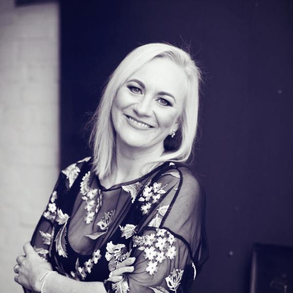 A professional black-and-white portrait of Marlene van Rooyen. She is smiling warmly, with her arms crossed in a confident yet approachable pose. Marlene is wearing a floral-patterned sheer top, and the neutral background emphasizes her radiant and engaging demeanor.